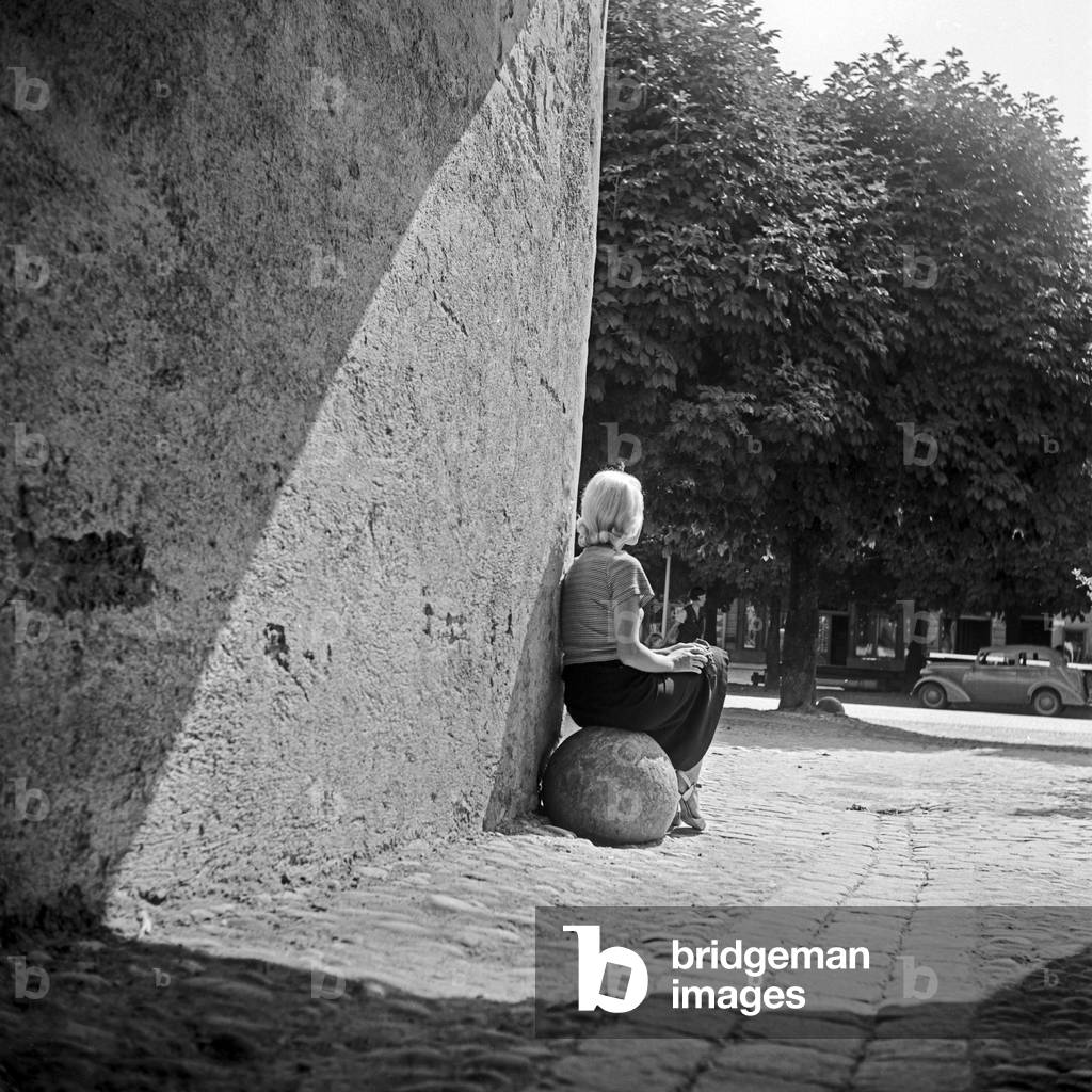 Immagine di Fotografia "Una giovane donna seduta su una palla di pietra ad una porta medievale della città", b